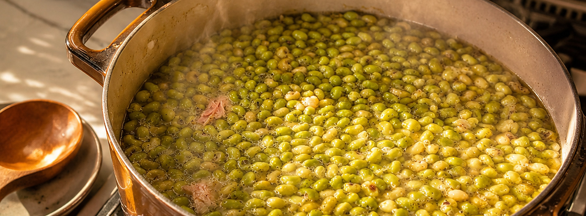 A copper pot of field peas simmering on a stove, a piece of ham in the pot, a wooden spoon resting on the counter beside it
